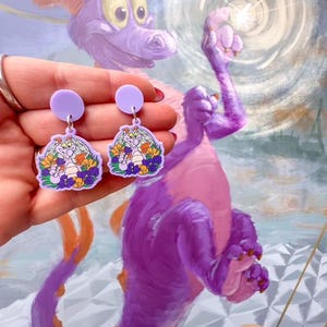 May include: Purple dangle earrings with a cartoon image of a purple dragon with yellow flowers. The dragon is wearing a crown and has a friendly expression.