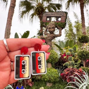 May include: A pair of red and black cassette tape earrings with the text "Awesome Mix Vol. 1" on each. The earrings are hanging from a person's ear. The background is a green, leafy garden with a large, wooden, cassette tape-shaped sculpture.