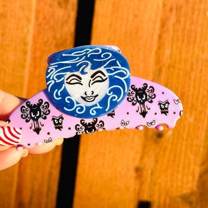 May include: A light pink hair claw clip with a blue circular design featuring a smiling face with curly hair. The clip has black and white haunted house and owl designs. A fun accessory for any hairstyle.