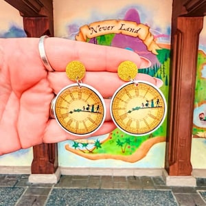 May include: A pair of round earrings featuring a clock face design with a silhouette of people. The earrings have yellow glittery tops and are held up in front of a mural with the words "Never Land".