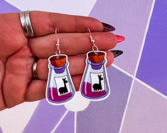 May include: A pair of silver earrings with purple potion bottles and a black llama silhouette. The bottles have a cork stopper and a white label.