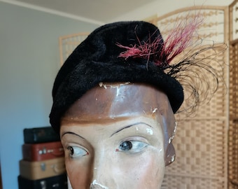 May include: A black velvet hat with a red feather accent. The hat is perched on a mannequin head with a light blue dress.