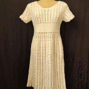 May include: A cream-coloured, short-sleeved crochet dress on a mannequin. The dress has a fitted bodice, a defined waist, and a flared skirt. The open crochet pattern gives a textured appearance. The dress is knee-length.