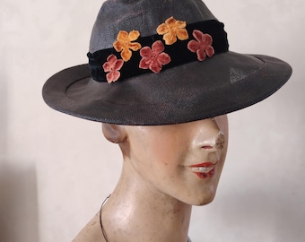 May include: A dark gray fedora hat with a wide brim. The hat features a black velvet band adorned with five velvet flower appliqués in shades of orange and red. The hat is displayed on a mannequin.