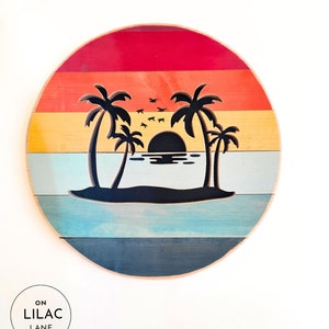 May include: A round wooden wall decor with a sunset scene featuring palm trees and a silhouette of a setting sun. The background is painted in a retro color palette of red, orange, yellow, blue, and green.