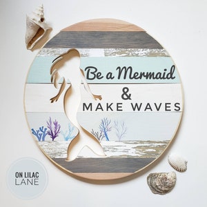 May include: A round wooden sign with a mermaid silhouette cut out. The sign has a distressed, weathered look with blue and gray paint. The text on the sign reads "Be a Mermaid & Make Waves".