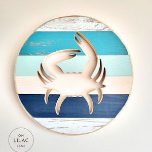 May include: A round wooden wall decor with a cutout of a crab in the center. The wood is painted in shades of blue, green, and white, creating a striped pattern. The crab is a natural wood color.