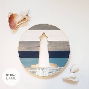 May include: A round wooden sign with a lighthouse silhouette cut out. The sign is painted in shades of blue, gray, and white, resembling a seascape. The text "ON LILAC LANE" is printed in white on a black circle in the lower left corner.