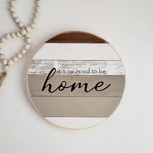 May include: Round wooden sign with a distressed white and gray paint finish. The sign has the text "It's so good to be home" in black lettering.