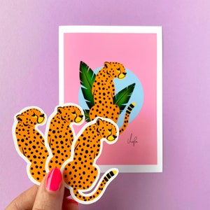 Könnte beinhalten: Ein rosa Hintergrund mit einer Gepardenillustration und drei Gepardenaufklebern. Die Illustration zeigt einen Geparden mit schwarzen Flecken, grünen Blättern und einem blauen Kreis. Die Aufkleber sind orange mit schwarzen Flecken.
