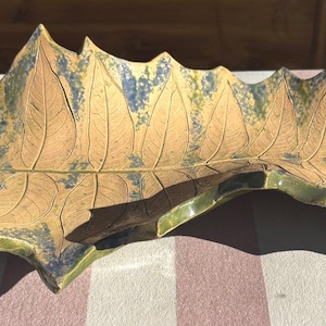 Op de afbeelding: Een keramische blad-vormige schaal met een getextureerd oppervlak. De schaal heeft een lichtbruine basiskleur met donkerdere bruine aderen en accenten van blauw en groen. De randen zijn geschulpt en de schaal ligt op een gestreept oppervlak.