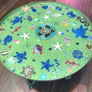 May include: A round, light green metal tray table featuring a central beaded sea turtle emblem surrounded by various marine life illustrations, including blue sea turtles, white starfish, colourful fish, and seashells, all accented with glitter.