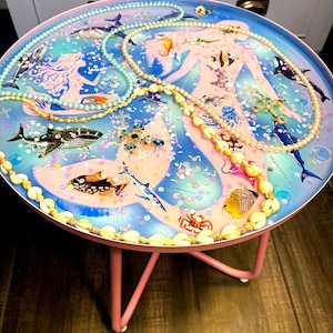 May include: A round side table with a mermaid and ocean-themed design. The table features a pink metal frame and a tabletop with a blue and pink illustration of mermaids, fish, and sharks. Several necklaces and jewelry pieces are arranged on the table.