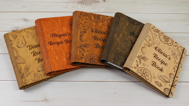 May include: Four wooden recipe books with different colored covers and personalized names. The covers feature a variety of kitchen-themed illustrations, including avocados, lemons, peppers, and pasta.