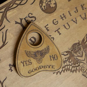 Op de afbeelding: Een houten Ouija-bord met een planchette. Het bord heeft gegraveerde letters, cijfers en symbolen, waaronder een uil en een wereldbol. De planchette heeft een uilontwerp en de woorden "YES", "NO" en "GOODBYE".