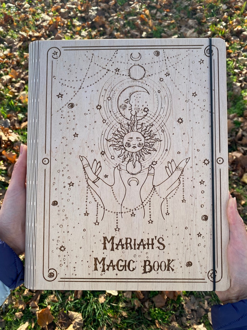 May include: A wooden journal with a celestial design, including a sun, moon, and stars. The journal has a black elastic closure and the text "MARIAH'S MAGIC BOOK" is engraved on the front.