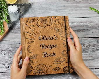 May include: A wooden recipe book with a black elastic closure. The cover is decorated with a laser-engraved design of various food items, including a lemon, pepper, and cheese. The text "Olivia's Recipe Book" is engraved on the cover.