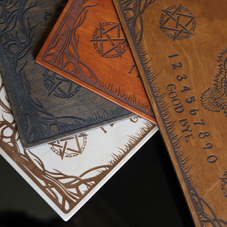 Op de afbeelding: Close-up van verschillende houten Ouija-borden in verschillende kleuren, waaronder bruin, grijs en wit. Elk bord heeft ingewikkelde gegraveerde ontwerpen van bomen, pentagrammen en cijfers. E&eacute;n bord toont de woorden "GOOD BYE".