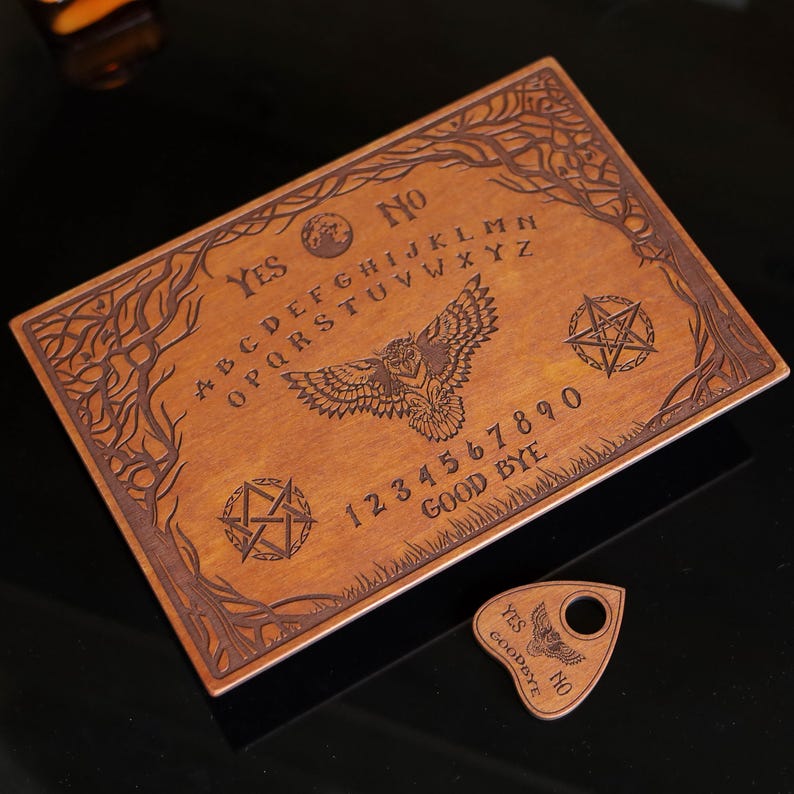 Op de afbeelding: Een houten Ouija-bord met gegraveerde letters, cijfers en symbolen. Het bord heeft een uilontwerp en de woorden "Ja" en "Nee". Een bijpassende planchette is inbegrepen. Het bord is bruin met een lichte houtnerf.