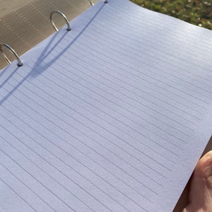 May include: A white notebook with lined pages and a silver binder. The notebook is open and lying flat on a wooden surface.