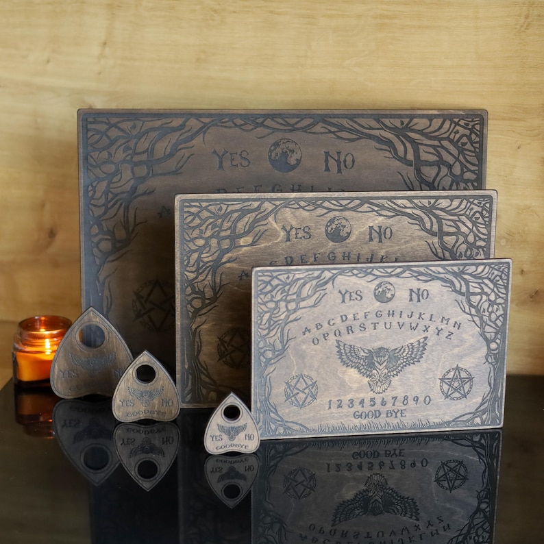 Op de afbeelding: Een verzameling houten Ouija-borden en planchette-aanwijzers. De borden zijn gegraveerd met letters, cijfers en de woorden "Ja" en "Nee". De planchette-aanwijzers hebben een rond kijkgaatje. De borden hebben een uil- en takontwerp.