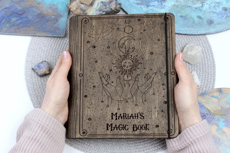 May include: A wooden journal with a celestial design, featuring a sun, moon, and stars. The journal has a black elastic closure and the text "MARIAH'S MAGIC BOOK" is engraved on the front.