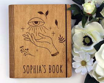 May include: A wooden journal with a brown wood cover and a black elastic band closure. The cover has an engraved design of an eye with rays of light, a hand, and a few small floral designs. The text "SOPHIA'S BOOK" is engraved at the bottom of the cover.