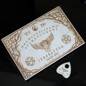 Op de afbeelding: Een houten Ouija-bord met gegraveerde letters, cijfers en symbolen. Het bord heeft een uil, pentagrammen en boomtakontwerpen. Inclusief een planchette met de woorden "Ja", "Nee" en "Vaarwel".