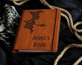 May include: A rectangular wooden book with a smooth, medium-brown finish. The front cover features a dark, laser-engraved silhouette of a witch flying on a broomstick, positioned above the engraved text "Anna's Book" in a simple, dark font.