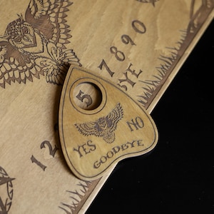 Op de afbeelding: Een houten Ouija-bord met gegraveerde cijfers, letters en een uilontwerp. De planchette bevat de woorden "JA", "NEE" en "GEDAG", samen met een uilillustratie en het nummer "5".