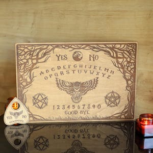 Op de afbeelding: Een houten Ouija-bord met gegraveerde letters, cijfers en symbolen. Het bord toont een uil, pentagrammen en de woorden "Ja", "Nee" en "Vaarwel". Een planchette en kaarsen zijn ook zichtbaar.