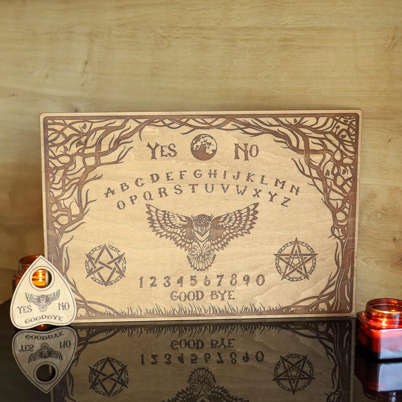 Op de afbeelding: Een houten Ouija-bord met gegraveerde letters, cijfers en symbolen. Het bord toont een uil, pentagrammen en de woorden "Ja", "Nee" en "Vaarwel". Een planchette en kaarsen zijn ook zichtbaar.