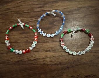 May include: Three beaded bracelets with holiday-themed messages. One bracelet says "MERRY CHRISTMAS" in red, green, and white beads. Another says "LET IT SNOW" in blue and white beads. The third says "PEACE ON EARTH" in green and red beads.