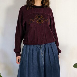 Könnte beinhalten: Ein maroonfarbener Pullover mit einem braunen und goldenen geometrischen Muster auf der Vorderseite, kombiniert mit einem blauen Jeans-Maxirock.