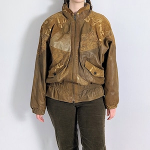 Puede incluir: Una chaqueta de cuero marrón con un diseño estampado en dorado. La chaqueta tiene cierre de cremallera y cuello alto.