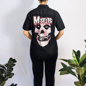 以下が含まれることがあります： 背面にミッフィーズの骸骨のグラフィックが施された、黒い半袖ボタンダウンシャツ。骸骨は白で、赤い塗料が垂れており、バンド名は骸骨の上に白で書かれています。