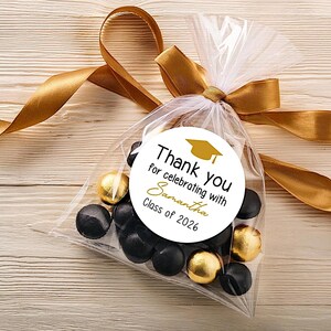 May include: A clear plastic favor bag filled with black and gold foil-wrapped candies, tied with a gold satin ribbon. A circular white sticker features a gold graduation cap icon and the text: Thank you for celebrating with Samantha Class of 2026.