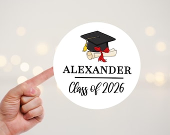 Pode incluir: Um autocolante redondo branco com um barrete de finalista preto com borla amarela e um diploma enrolado atado com uma fita vermelha. Abaixo da ilustração, o texto diz ALEXANDER em letras maiúsculas pretas e Class of 2026 em cursivo.