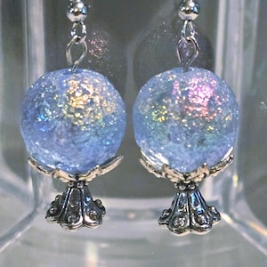 May include: A pair of earrings featuring light blue, glittery orbs with iridescent accents. The orbs are mounted on ornate silver-colored bases and suspended from silver-colored fish hook ear wires. The earrings are displayed in a clear, plastic container.