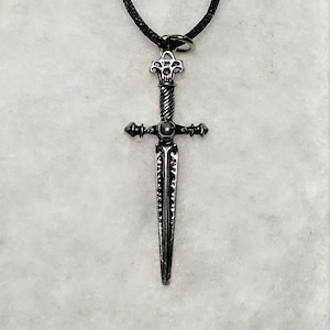 May include: A silver dagger pendant on a black cord necklace. The dagger has a detailed design with a twisted handle and a pointed tip.