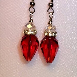 May include: A pair of dangle earrings featuring red, faceted teardrop-shaped beads. Each earring has a clear, faceted bead above the red bead, with gold-colored metal accents and ear wires. The earrings are displayed against a white background.
