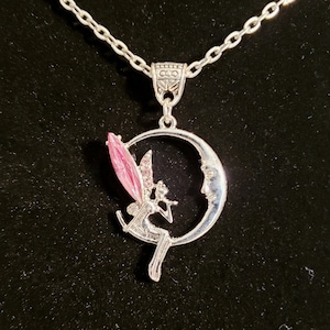 May include: A silver necklace with a crescent moon pendant featuring a fairy with pink wings sitting on the moon.