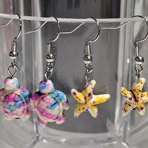 May include: Four pairs of dangle earrings. Two pairs feature turtle shapes with pink, blue, and white patterns. The other two pairs have starfish shapes in yellow with brown spots. All earrings have silver-colored hooks.