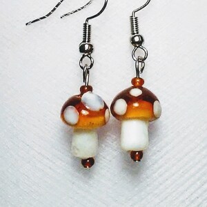 May include: A pair of mushroom-shaped earrings. The earrings feature a brown cap with white spots and a white stem. The earrings are suspended from silver-colored fish hook ear wires. The earrings are a whimsical accessory.