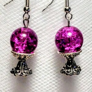 May include: A pair of dangle earrings with a silver base and a round, purple glass bead. The beads have a sparkly, iridescent finish.