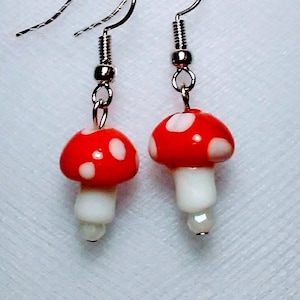 May include: A pair of silver dangle earrings featuring red and white mushroom charms. The mushroom charms have white polka dots on the red cap and a white stem.