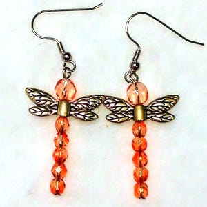 May include: A pair of dragonfly earrings. Each earring features a silver-colored metal dragonfly body with orange beaded accents. The wings are detailed with a textured pattern, and the body is adorned with a series of orange beads. The earrings have silver-colored ear wires.