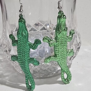 May include: A pair of green alligator-shaped earrings. The earrings are made of a shiny, metallic material and feature detailed scales and limbs. The earrings hang from silver-coloured fish hook ear wires. The earrings are displayed in front of a clear glass.