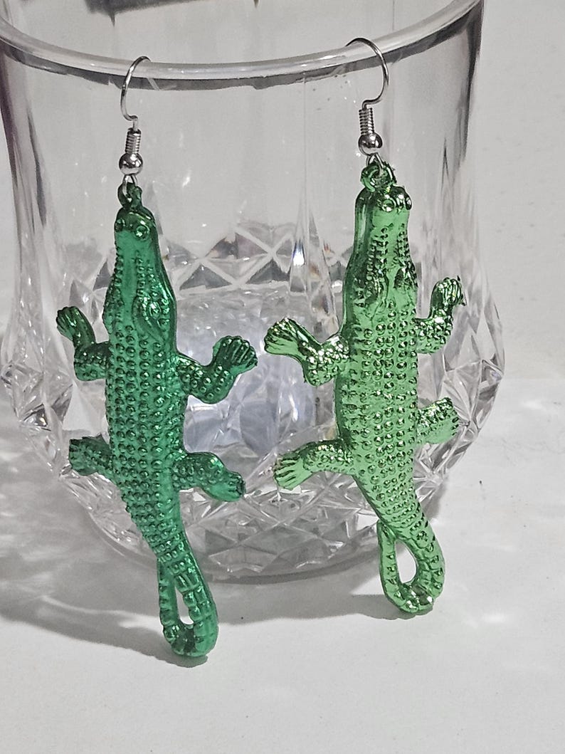 May include: A pair of green alligator-shaped earrings. The earrings are made of a shiny, metallic material and feature detailed scales and limbs. The earrings hang from silver-coloured fish hook ear wires. The earrings are displayed in front of a clear glass.