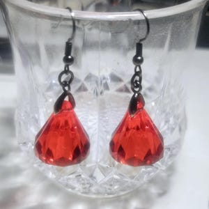May include: A pair of dangle earrings with red faceted teardrop-shaped crystals and black metal hooks.